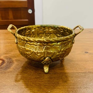 Peut inclure: Un bol rond en laiton avec une surface texturée ornée de motifs de feuilles et de vignes en relief. Le bol possède deux petites anses latérales, un bord torsadé en forme de corde et repose sur trois pieds courts et décoratifs en métal doré.
