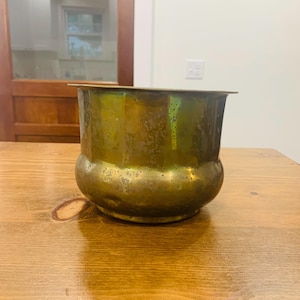 Pode incluir: Um vaso de latão manchado com uma base arredondada e uma borda ligeiramente alargada. O vaso tem uma aparência desgastada, com variações de cor de ouro a verde. Está sobre uma superfície de madeira.
