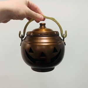 May include: A round, copper-colored metal pot with a lid and a brass-toned handle. The front of the pot features a cutout design resembling a jack-o'-lantern face with two triangular eyes and a wide, curved mouth with a single square tooth.