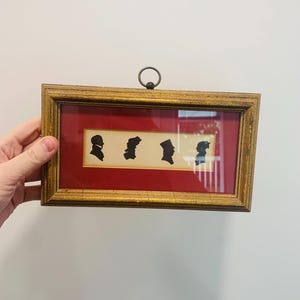 May include: A rectangular gold-toned frame featuring a deep red mat and a cream-colored insert. Inside the insert are four black silhouette profiles of heads facing right. The frame includes a small circular metal ring at the top for hanging.