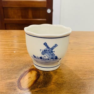 May include: A small, cream-coloured ceramic cup featuring a blue windmill design. The cup has a scalloped rim and a blue line around the top and base. The windmill image is detailed with blue lines.