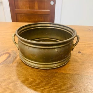 Pode incluir: Um vaso redondo de latão com duas alças. O vaso tem um aspeto desgastado e envelhecido.