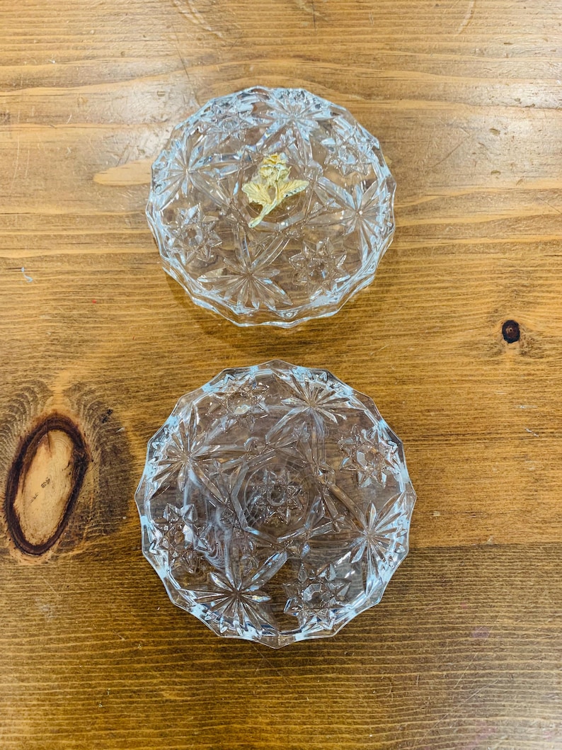 May include: Two clear glass trinket boxes with a starburst pattern. The top box has a gold rose design on the lid.
