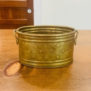 Peut inclure: Une bassine ovale en laiton présentant une surface texturée avec des motifs d'étoiles en relief. Le récipient possède un design nervuré horizontal sur les bords supérieur et inférieur, ainsi que deux petites anses incurvées sur les côtés.