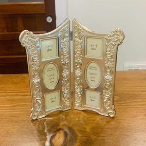 May include: A silver-colored hinged double photo frame featuring ornate scrollwork borders. Each panel contains three openings: two square slots for 1.5 x 1.5 inch photos and one central oval slot for a 1.5 x 2 inch photo, labeled Godinger Silver Art.