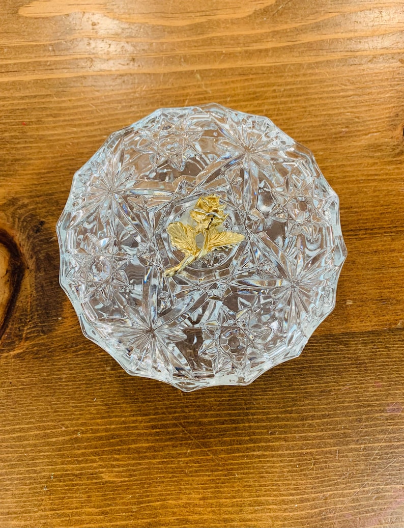 May include: A clear glass trinket box with a cut glass design and a gold rose accent.