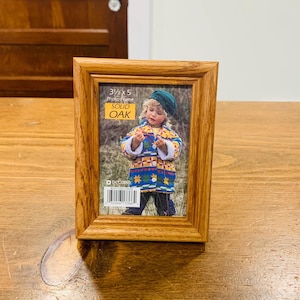 May include: A rectangular solid oak photo frame, displaying a 3.5 x 5 inch image. The frame has a natural wood grain finish and features the text "3 1/2 x 5 Photo Frame SOLID OAK". The image shows a child wearing a colorful coat.