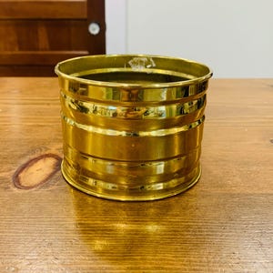 Könnte beinhalten: Ein zylindrischer, goldfarbener Metallbehälter mit horizontalen Bändern. Der Behälter besteht aus einem glänzenden, reflektierenden Material und steht auf einer Holzoberfläche. Der Behälter ist leer und scheint ein dekorativer Gegenstand zu sein.