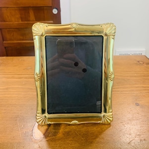 May include: A gold-coloured picture frame with an ornate, scalloped design. The frame has a rectangular opening with a dark interior, likely for a photograph. The frame is sitting on a wooden surface.