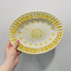May include: Oval serving platter with a yellow and white floral design. The platter has a scalloped edge and a large flower pattern in the centre. The border features a repeating floral and leaf pattern. The colour palette is primarily yellow and white.