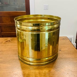 Könnte beinhalten: Ein zylindrischer Übertopf aus Messing mit polierter Metalloberfläche und vertikalen Rillen an den Seiten. Der Behälter hat eine weite Öffnung mit verstärktem Rand und einen flachen Boden, wobei leichte Gebrauchsspuren und eine natürliche Patina sichtbar sind.