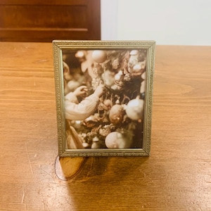 May include: A rectangular picture frame with a metallic gold finish and a textured, repeating geometric pattern along the border. The frame contains a photograph showing a person decorating a Christmas tree with various round ornaments.