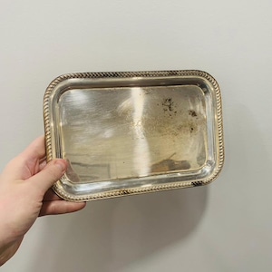 May include: A rectangular silver-toned metal tray featuring a polished, reflective surface and a decorative rope-style border along the raised edges. The tray shows signs of natural oxidation and minor surface wear across the center.