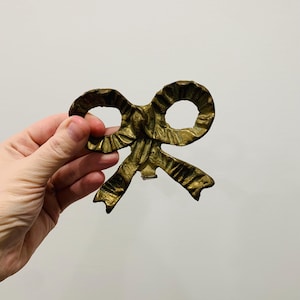 Peut inclure: Un objet décoratif vintage en forme de nœud papillon en laiton. Le nœud présente une surface texturée et détaillée avec une teinte dorée chaude. Le design comprend deux grandes boucles et des extrémités de ruban fluides, suggérant un style ornemental classique.