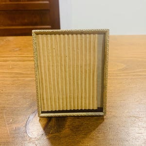 May include: A rectangular picture frame with a thin, metallic gold-toned border featuring a repeating triangular pattern. The interior of the frame contains a light beige, vertically ribbed paper insert that covers the majority of the display area.