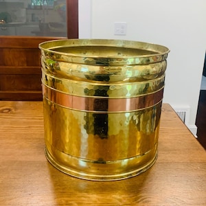 Puede incluir: Una jardinera cilíndrica de latón con textura martillada y una banda de cobre. La jardinera tiene una parte superior ancha y abierta y está sobre una mesa de madera. El latón tiene una superficie brillante y reflectante.