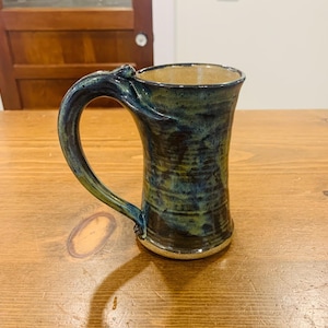 Könnte beinhalten: Handgefertigte Keramiktasse mit einzigartigem Griffdesign. Die Tasse hat eine dunkelblaue und grüne Glasur mit strukturierter Oberfläche. Innen und am Boden ist sie hellbraun. Diese Steinzeugtasse ist perfekt für Kaffee oder Tee.