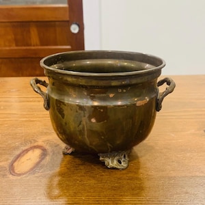 Pode incluir: Um pequeno pote redondo de metal em tom de bronze escuro com duas alças laterais e três pés decorativos ornamentados. A superfície do metal apresenta sinais de desgaste e oxidação, com manchas visíveis de pátina em tom cobre claro.