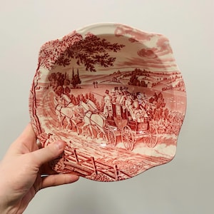 May include: A decorative, irregularly shaped plate with a red and white transferware design. The plate features a detailed scene of a horse-drawn carriage on a road, with trees and a fence. The plate is held by a hand.