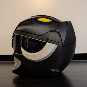 Op de afbeelding: Een zwarte plastic helm met een zilveren vizier en een gele streep aan de bovenkant. De helm heeft een getextureerd oppervlak en een zilveren ontwerp aan de voorkant.
