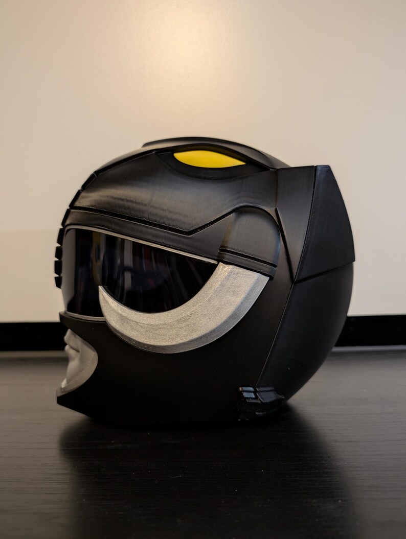 Op de afbeelding: Een zwarte plastic helm met een zilveren vizier en een gele streep aan de bovenkant. De helm heeft een getextureerd oppervlak en een zilveren ontwerp aan de voorkant.