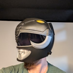 Op de afbeelding: Een zwarte en zilveren Power Rangers helm met een gele vizier. De helm heeft een zilveren gezichtsplaat met een mond en neus.