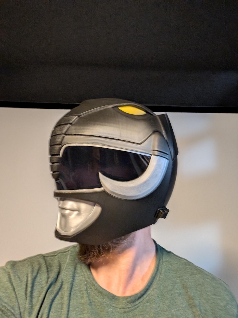 Op de afbeelding: Een zwarte en zilveren Power Rangers helm met een gele vizier. De helm heeft een zilveren gezichtsplaat met een mond en neus.