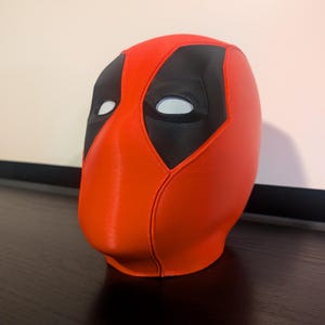 Puede incluir: Una m&aacute;scara de Deadpool roja y negra. La m&aacute;scara es principalmente roja con detalles negros alrededor de los ojos y una secci&oacute;n superior negra. Las aberturas de los ojos son blancas con un material de malla. La m&aacute;scara est&aacute; sobre una superficie oscura.