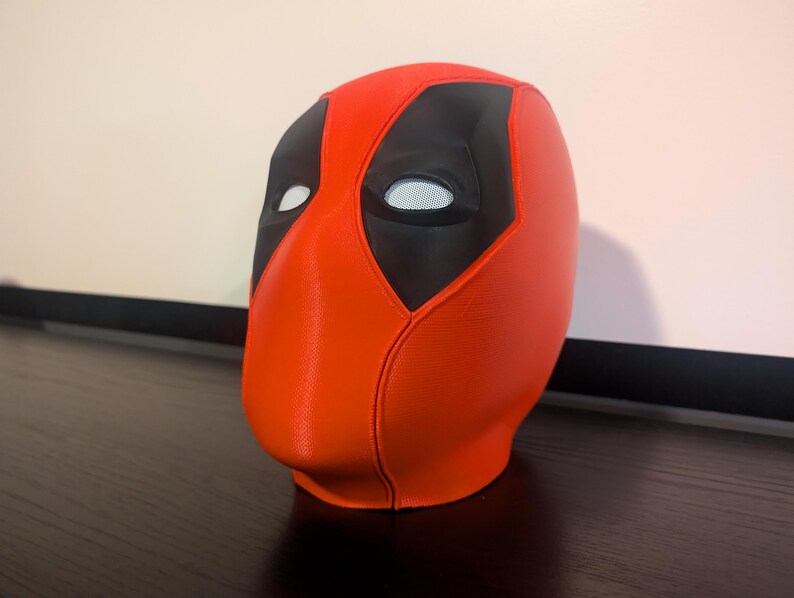 Puede incluir: Una m&aacute;scara de Deadpool roja y negra. La m&aacute;scara es principalmente roja con detalles negros alrededor de los ojos y una secci&oacute;n superior negra. Las aberturas de los ojos son blancas con un material de malla. La m&aacute;scara est&aacute; sobre una superficie oscura.