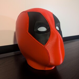 Puede incluir: Una m&aacute;scara de Deadpool roja y negra. La m&aacute;scara es principalmente roja con detalles negros alrededor de los ojos y la frente. Las aberturas de los ojos tienen un material de malla. La m&aacute;scara est&aacute; sobre una superficie oscura.