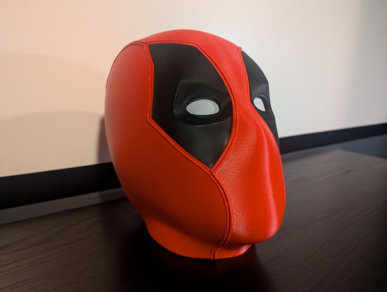 Puede incluir: Una m&aacute;scara de Deadpool roja y negra. La m&aacute;scara es principalmente roja con detalles negros alrededor de los ojos y la frente. Las aberturas de los ojos tienen un material de malla. La m&aacute;scara est&aacute; sobre una superficie oscura.