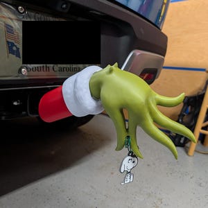 May include: A green, long-fingered hand with a red and white cuff attached to a vehicle hitch receiver. The hand holds a small, white, black-outlined cartoon dog charm dangling from a green ring. The object is positioned below a license plate.