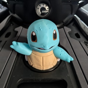 Puede incluir: Una figura del personaje Squirtle en color azul claro y beige con grandes ojos oscuros, cabeza redondeada y caparazón color canela. La figura está colocada en una abertura circular sobre una superficie gris oscuro con un logo BRP circular.
