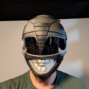 Op de afbeelding: Een zwarte en zilveren Power Rangers helm met een vizier. De helm heeft een getextureerd oppervlak en een zilveren mondbeschermer.