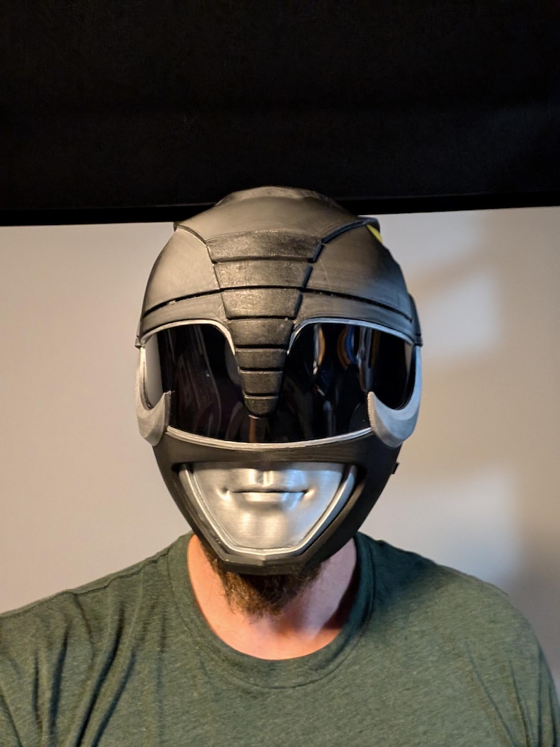 Op de afbeelding: Een zwarte en zilveren Power Rangers helm met een vizier. De helm heeft een getextureerd oppervlak en een zilveren mondbeschermer.