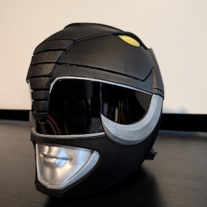 Op de afbeelding: Zwarte Power Rangers helm met zilveren accenten en een geel vizier. De helm is gemaakt van plastic en heeft een getextureerd oppervlak.