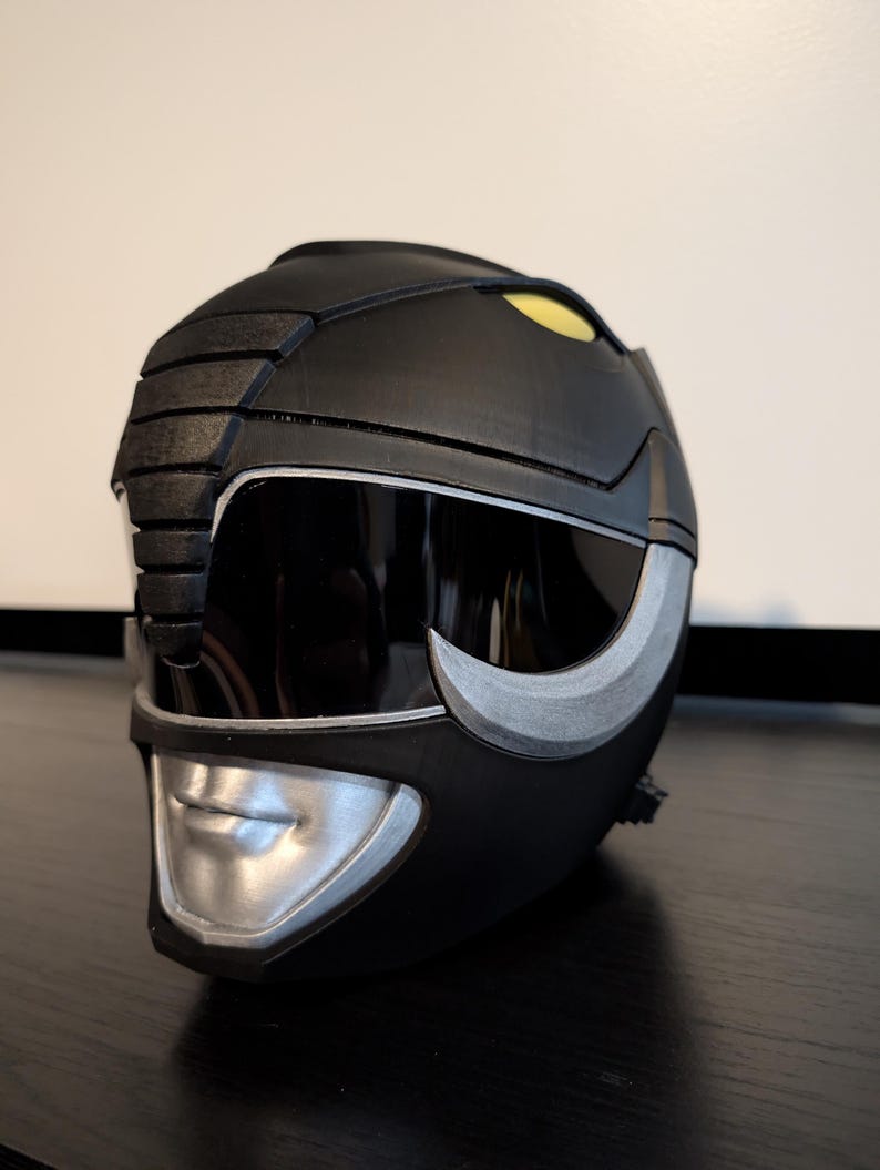 Op de afbeelding: Zwarte Power Rangers helm met zilveren accenten en een geel vizier. De helm is gemaakt van plastic en heeft een getextureerd oppervlak.