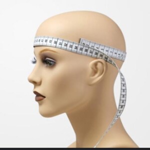 Puede incluir: Una cabeza de maniqu&iacute; con una cinta m&eacute;trica blanca envuelta alrededor de la cabeza. La cinta est&aacute; marcada en cent&iacute;metros. El maniqu&iacute; tiene un tono de piel neutro, con maquillaje aplicado en los ojos y los labios.