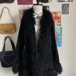 May include: A black coat with a faux fur collar, cuffs, and trim, displayed on a mannequin. The coat has a suede-like texture and a classic silhouette. A black necklace is visible. The background includes posters and handbags.