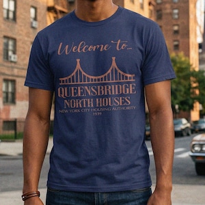 May include: A navy blue t-shirt with the text "Welcome to..." above a bridge graphic and "Queensbridge North Houses" below. The shirt also includes "New York City Housing Authority 1939".