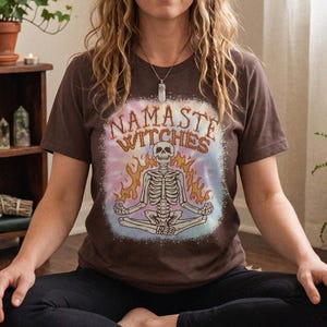 May include: A brown short-sleeve t-shirt featuring a graphic of a skeleton sitting in a lotus position surrounded by flames. The text "NAMASTE WITCHES" is printed in an arched font above the skeleton, set against a light blue tie-dye background.