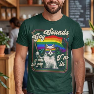 May include: A dark green t-shirt featuring a graphic of a white cat wearing rainbow-coloured sunglasses and holding a rainbow flag. The text reads "Sounds Gay I'm in" and "a tini bit gay" in a retro font, surrounded by colourful concentric square borders.