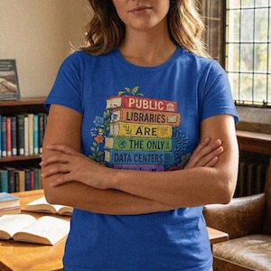 May include: A royal blue short-sleeved t-shirt featuring a graphic of a stack of colorful books. The text on the book spines reads: PUBLIC LIBRARIES ARE THE ONLY DATA CENTERS. The design is accented with small illustrations of green leaves and flowers.