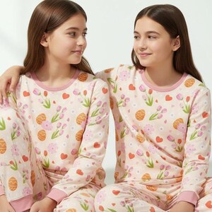 Puede incluir: Dos personas con pijamas a juego. Los pijamas son de color rosa claro con un estampado de huevos de Pascua, flores y corazones. La parte superior de manga larga y los pantalones tienen ribetes rosas. Las personas est&aacute;n sentadas juntas.