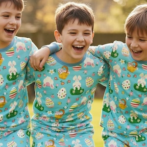 May include: Three children wearing matching light blue pajamas with an Easter-themed pattern of eggs, bunnies, and baskets. They are smiling and embracing each other outdoors in a grassy area.