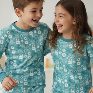 May include: Two children wearing matching teal pajamas with an Easter-themed pattern of eggs, bunnies, and flowers. The long-sleeved tops have a crew neck and cuffs in a darker teal. The children are smiling and laughing.