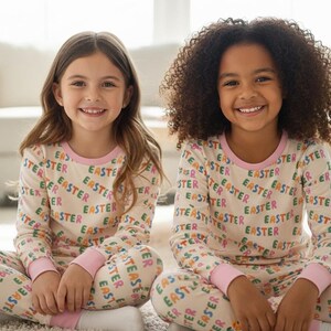 K&ouml;nnte beinhalten: Zwei junge M&auml;dchen tragen passende beige Pyjamas mit dem Wort "EASTER" in bunten Buchstaben. Die Pyjamas haben einen rosa Besatz an Ausschnitt und Manschetten. Sie sitzen auf einem wei&szlig;en Teppich und l&auml;cheln in die Kamera.