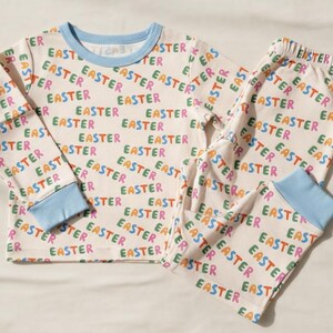 K&ouml;nnte beinhalten: Ein zweiteiliger Schlafanzug mit cremefarbenem Hintergrund und dem Wort "EASTER" in verschiedenen Farben. Das lang&auml;rmlige Oberteil und die Hose haben hellblaue Abschl&uuml;sse.