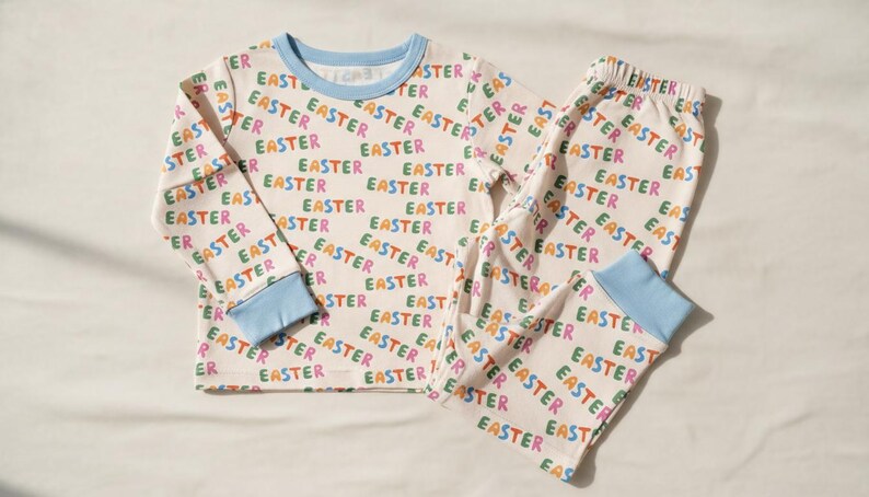 K&ouml;nnte beinhalten: Ein zweiteiliger Schlafanzug mit cremefarbenem Hintergrund und dem Wort "EASTER" in verschiedenen Farben. Das lang&auml;rmlige Oberteil und die Hose haben hellblaue Abschl&uuml;sse.