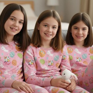 May include: Three individuals wearing matching pink pyjamas with Easter-themed patterns, including eggs and bunnies, are seated on a grey sofa. One person holds a white stuffed bunny. The pyjamas have long sleeves and trousers.